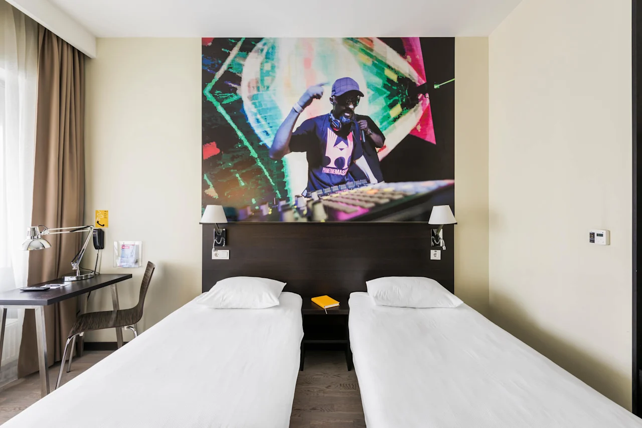 Two single beds in a hotel room, topped by a mural of a DJ performing, with a desk and a window adorned with brown curtains.