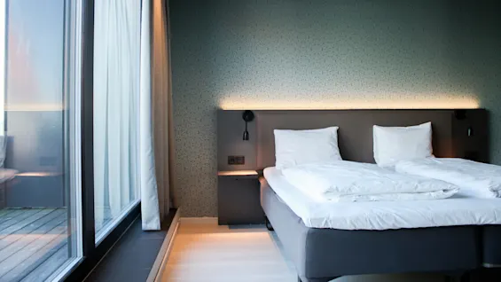 A neatly made double bed with white pillows and duvet stands against a patterned wall, illuminated by soft lighting. A large window with sheer curtains overlooks a deck.