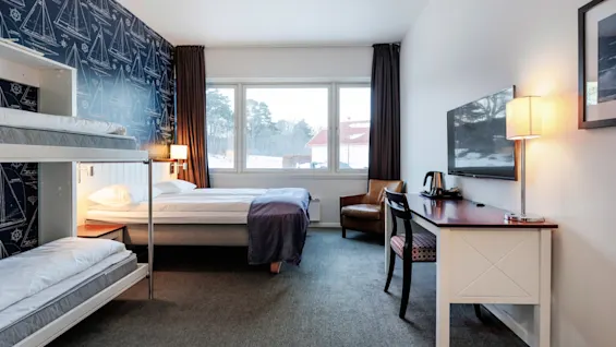 A hotel room with bunk beds and a double bed, features a nautical-themed blue wallpaper. A desk with a lamp, chair, and TV is on the right, facing a large window with snow-covered trees outside.