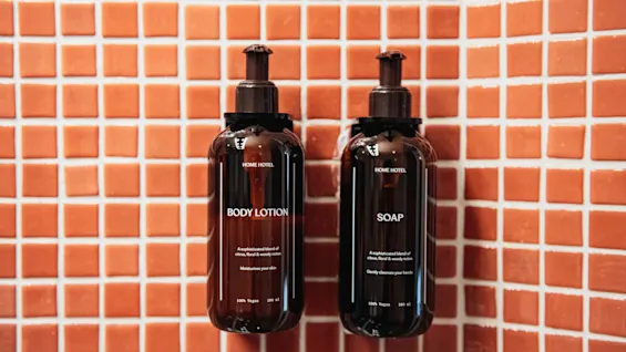 Home Hotel Uppsala body lotion and soap bottles on an orange tiled wall. The products offer a sophisticated blend of citrus, floral, and woody notes.