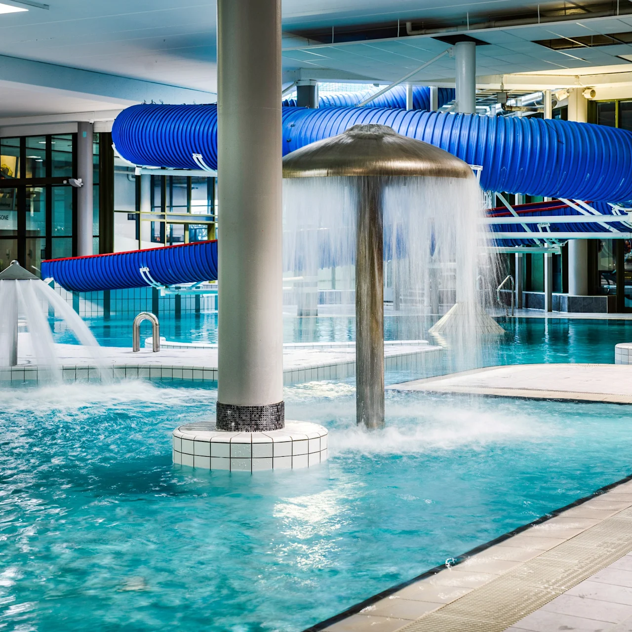 A large, indoor swimming pool with fountains spraying water, surrounded by tiled flooring and pillars. A blue spiral waterslide is in the background, coiled above the pool.