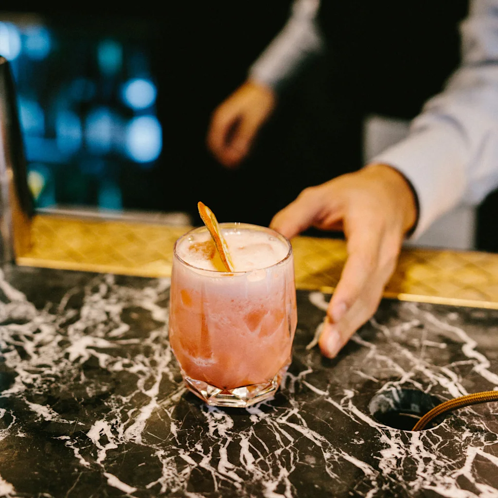 En bartenders hånd strekker seg etter en forfriskende rosa cocktail med garnityr på en stilig marmorbar.