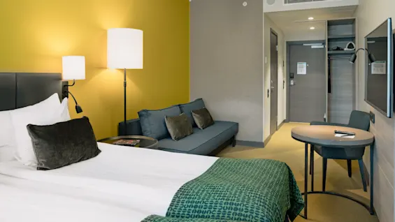 Ett hotellrum med en säng klädd i vitt och grönt linne, bredvid en blå soffa och ett bord med en stol, är upplyst av vägglampor mot en gul vägg.