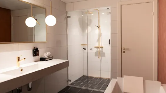 Home Hotel Grand Bodø: A modern bathroom with a double sink, a spacious shower, and a bathtub. Gold accents add a touch of luxury.