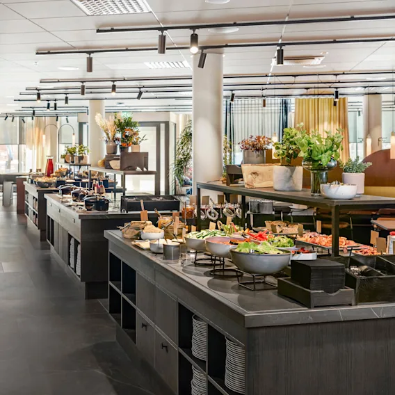 Buffet tables displaying assorted foods are arranged in a spacious, modern dining area. The setting includes ample lighting, decorative plants, and shelves of plates and cutlery.