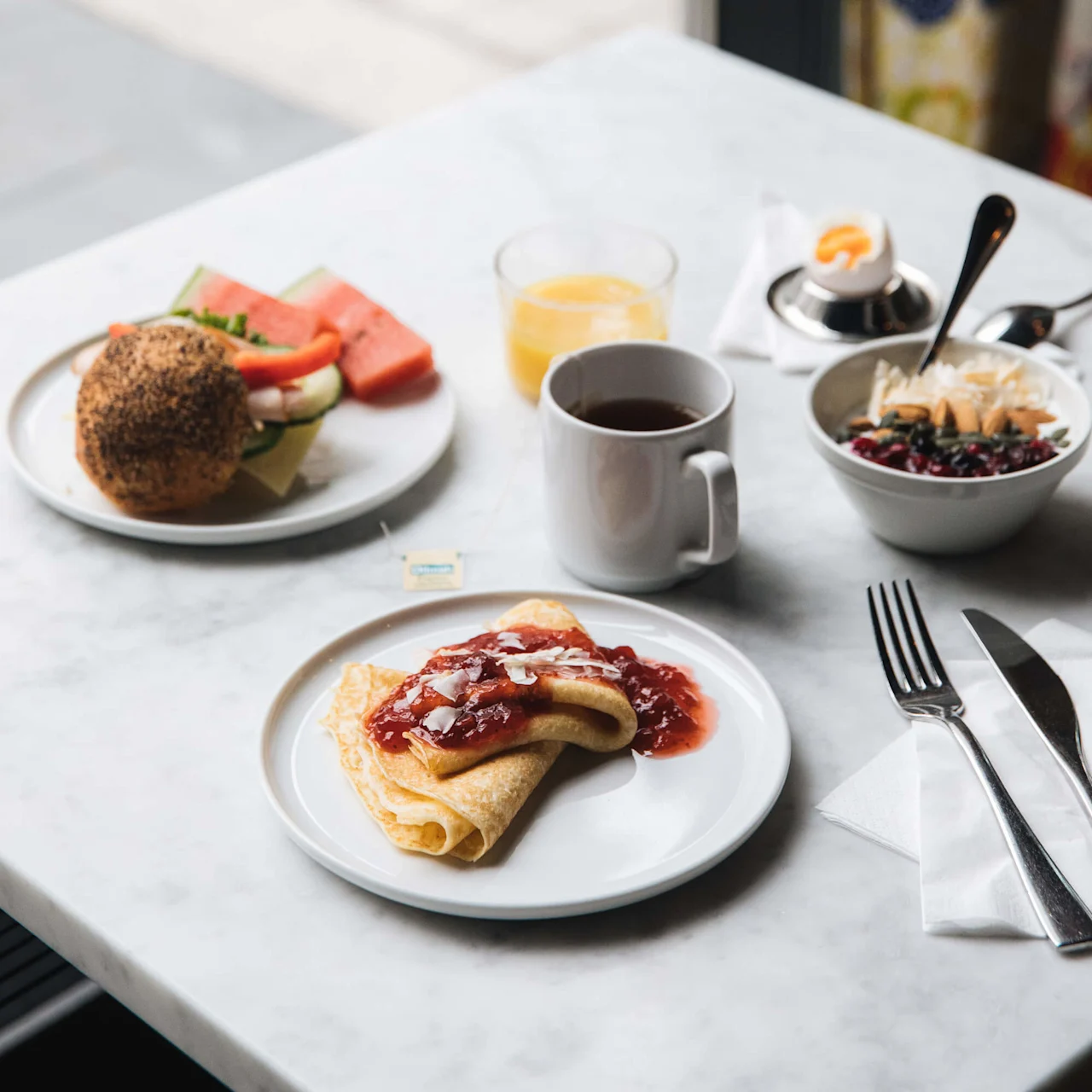Pancake with jam on a white plate rests on a marble table, accompanied by coffee, juice, a roll with fruit, and a bowl of yogurt near a window.