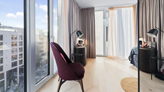 A purple chair faces a small desk with a black lamp in a bright room with large windows and beige curtains, overlooking an urban setting with buildings.