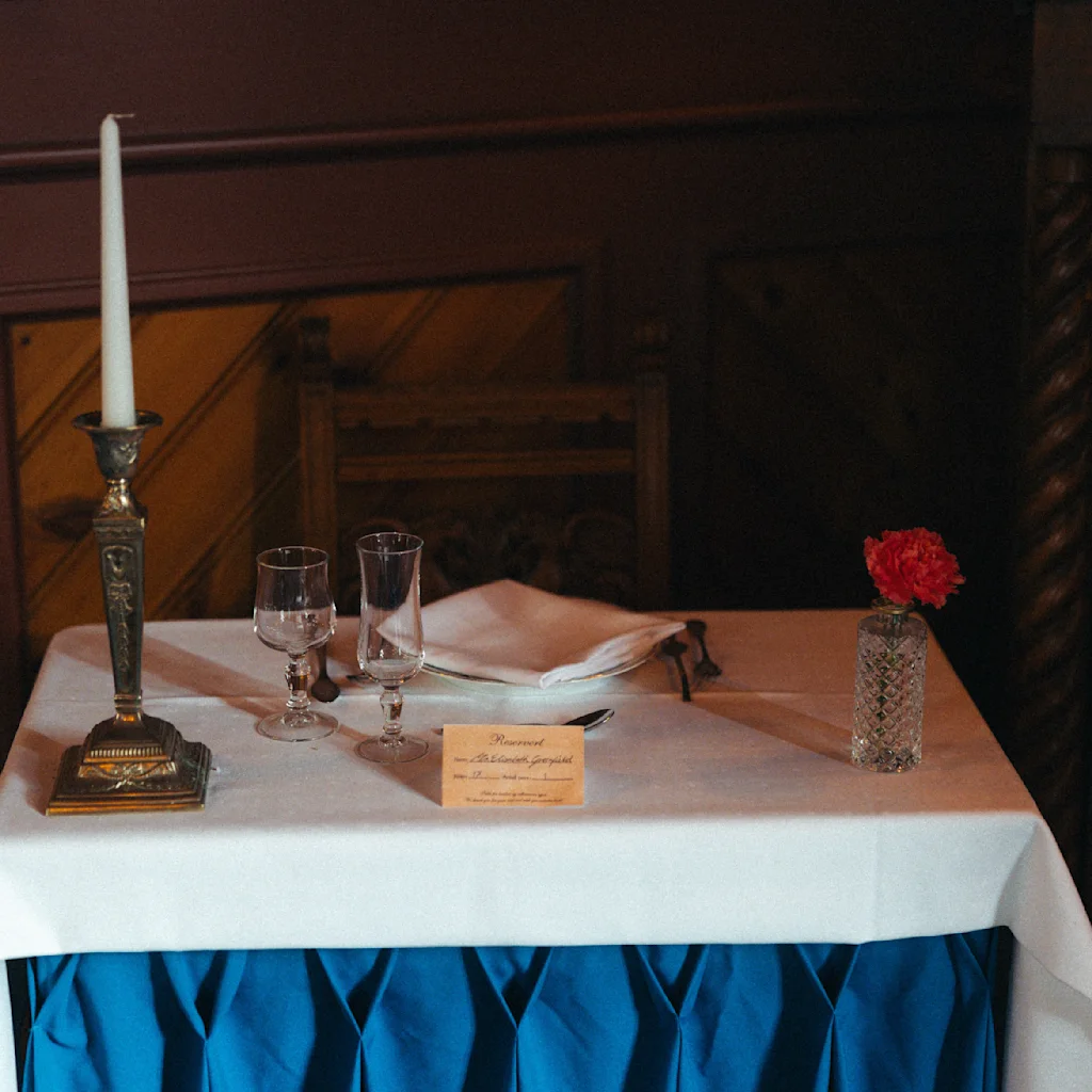 A cozy table setup at Dalen Hotel features a tall candlestick, elegant glasses, and a single red flower in a vase, creating an inviting atmosphere for dining. A small card adds a personal touch.