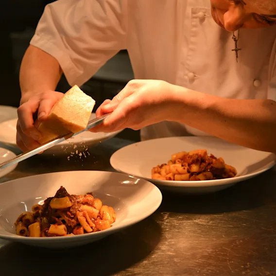 Chef grates cheese over pasta in a dimly lit kitchen, preparing multiple plates for serving.