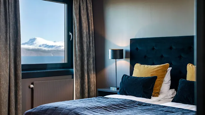 Cozy bed with dark blue and yellow pillows sits beside a window, offering a view of snow-capped mountains. Soft lighting complements the warm, inviting interior setting.