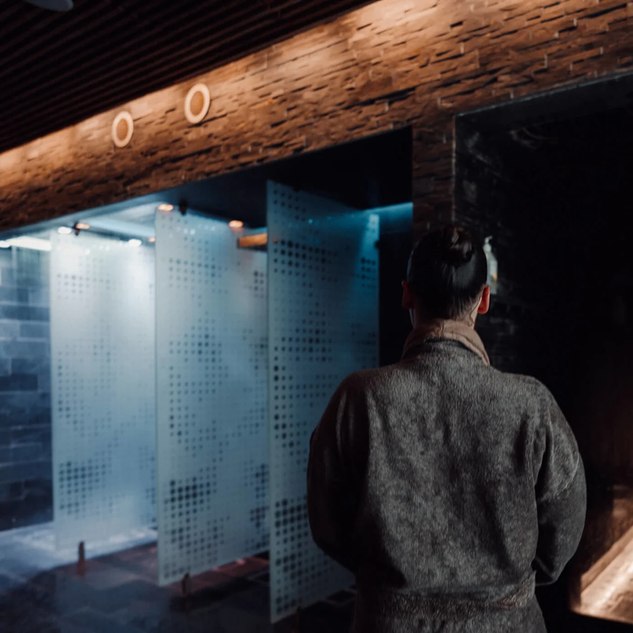 En person i en badekåpe står foran et moderne, mørkt spa-miljø med frostede glassvegger og dempet belysning.