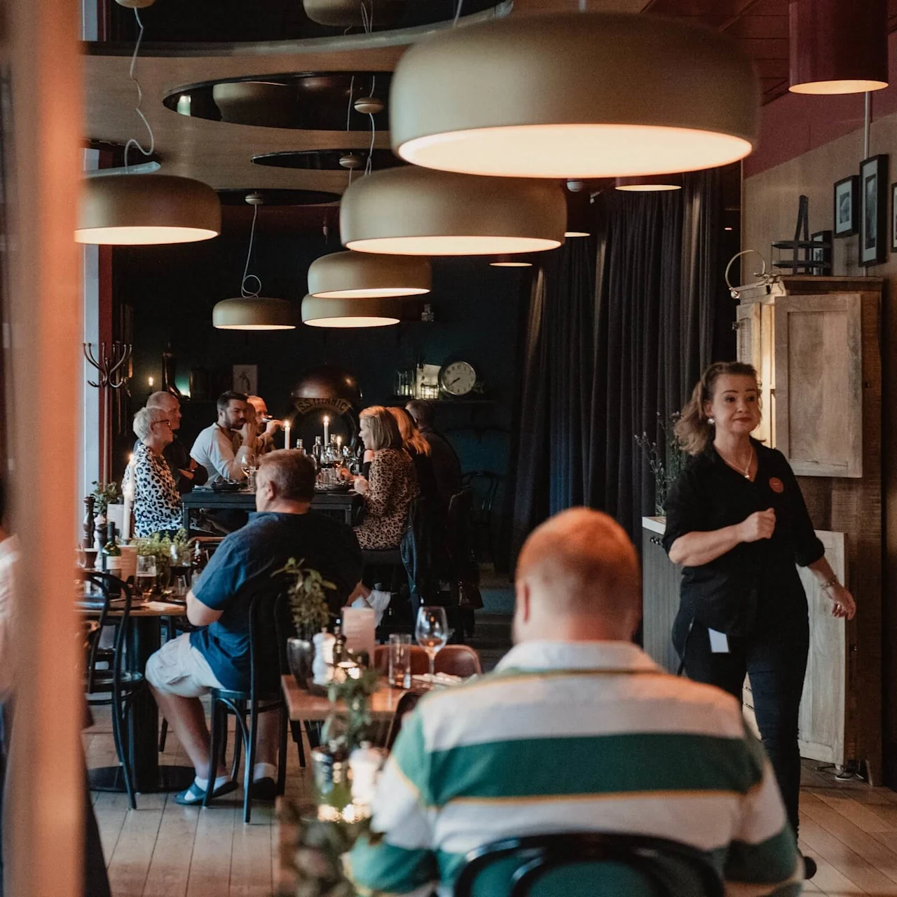 A group of people dine in a warmly lit restaurant with modern pendant lights. A waiter walks briskly, and framed pictures decorate the walls.