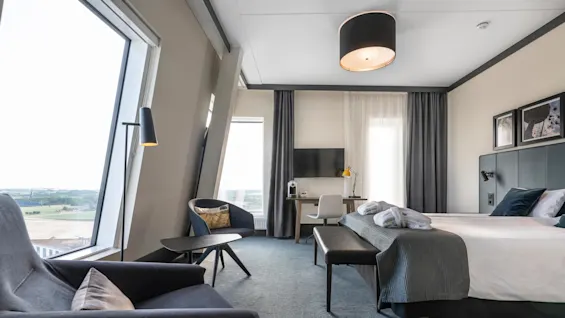 A modern hotel room features a large bed with pillows and a throw, a desk with a chair and lamp, and a seating area with armchairs near wide windows offering scenic views.