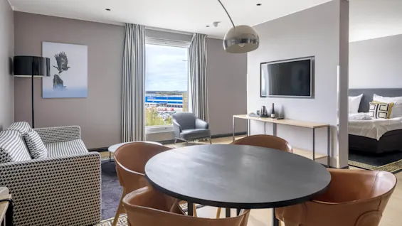 Circular table surrounded by leather chairs stands in a modern hotel room. A striped sofa, large TV, and bedside area are present. Window reveals an exterior view.