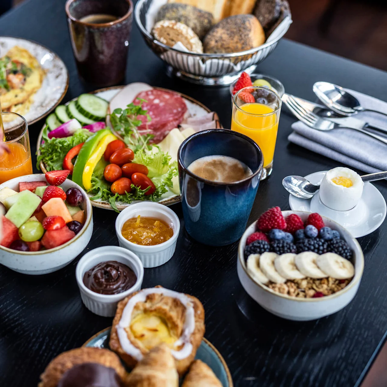 Ett bord visar en varierad frukost med frukter, bakverk, kött, sallad och drycker. En vas med blommor finns nära, vilket förhöjer den eleganta matsituationen.