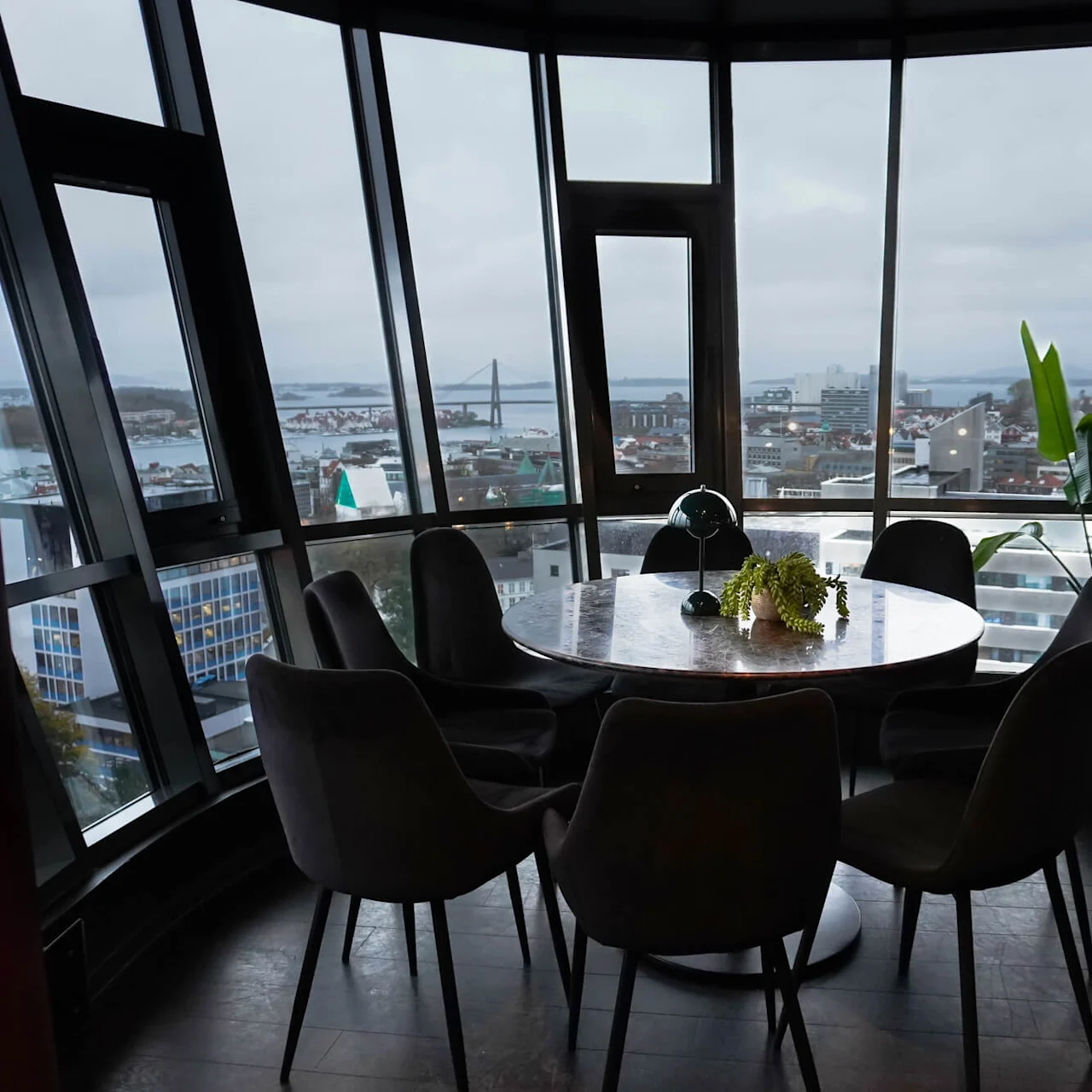 A round table with chairs, holding a plant and lamp, sits in a room with large windows offering a cityscape view, including a bridge and water.