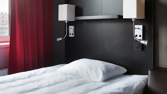 En säng med vita lakan och en kudde, inramad av röda gardiner och en mörk sänggavel med två moderna sänglampor som sträcker sig horisontellt.