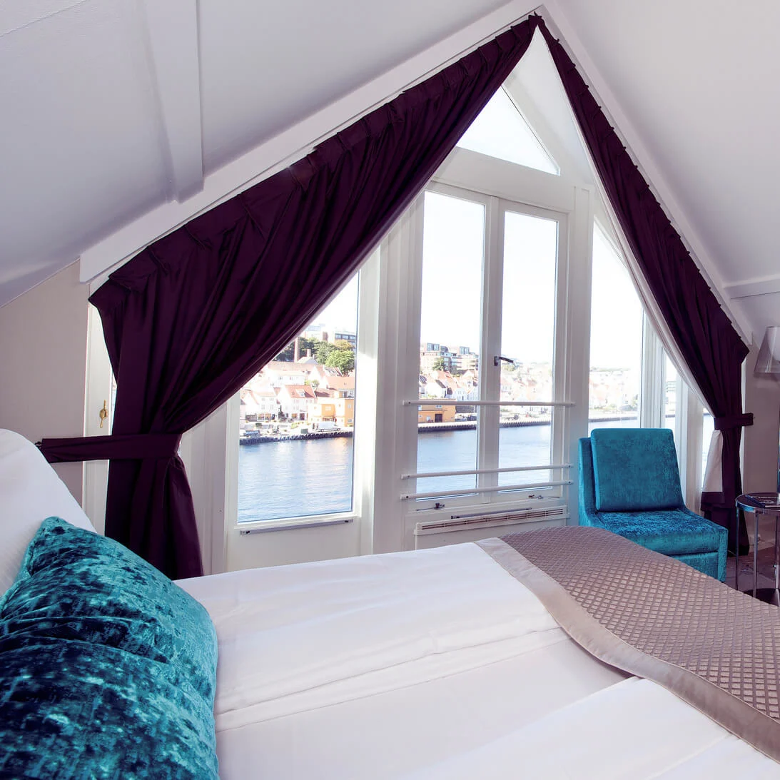A neat hotel room with a bay window overlooking a scenic waterfront, velvet chairs, and plush bedding against a calm, neutral decor.