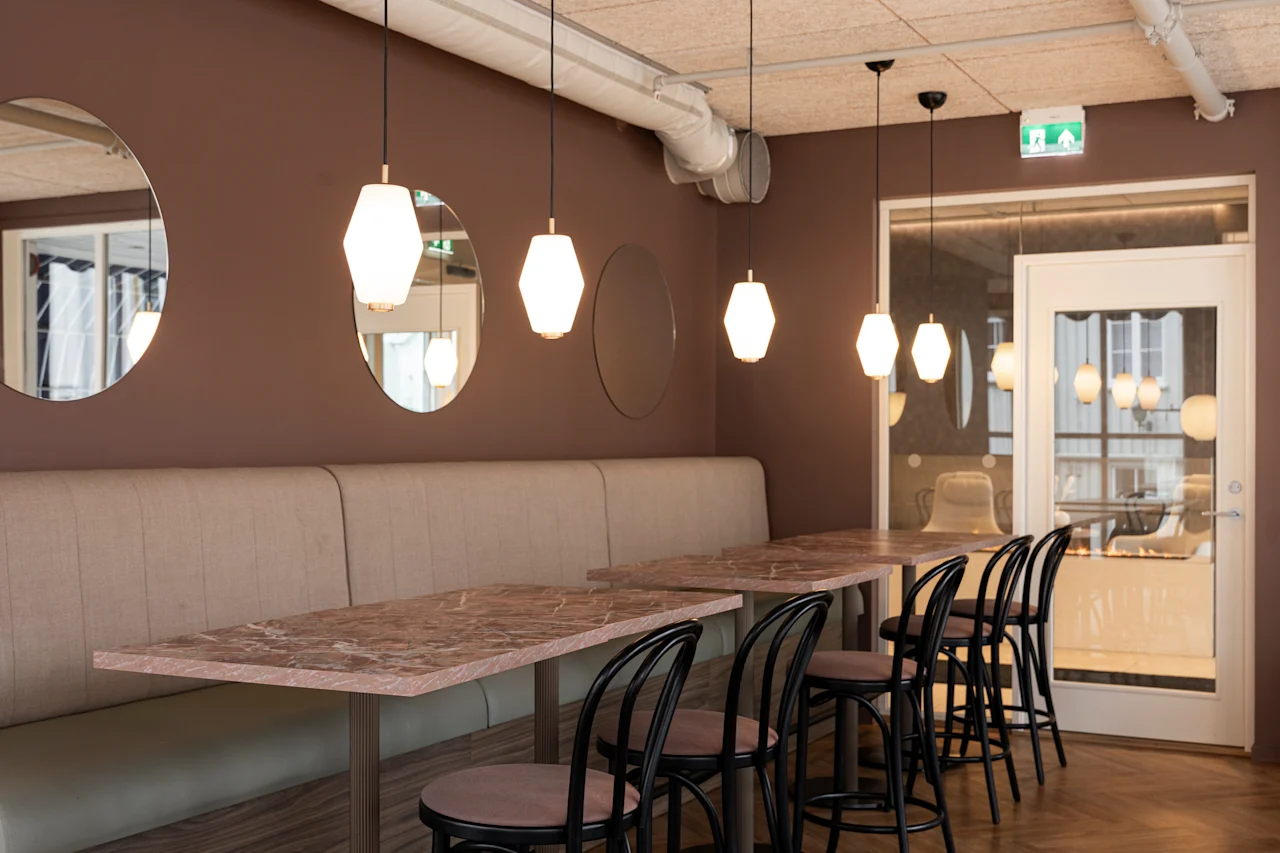 A modern cafe interior with marble-top tables, bench seating, and stylish pendant lights. An exit sign is visible above a glass door.