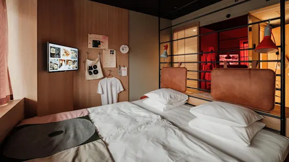 Bed with white linens against a wooden wall featuring mounted TV and various hanging items including a tote and a T-shirt. Warm lighting creates a cozy atmosphere.