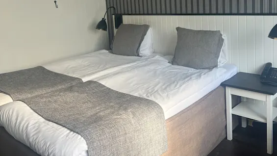 A neatly made bed with gray pillows and throws stands against a striped wall. A bedside table holds a black lamp and a phone in a softly lit room.
