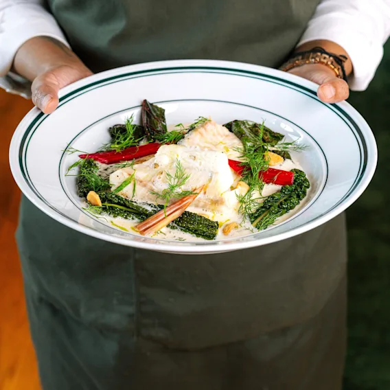 A person in a green apron holding a white plate with a delicious-looking meal of fish, green vegetables, and red peppers.