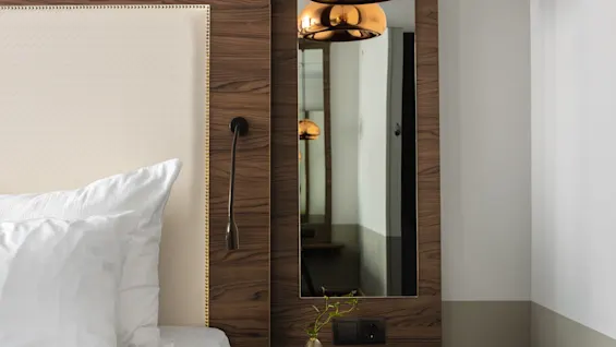 Ett mysigt hörn i hotellrummet med en prydlig säng med vita kuddar mot en trävägg. En stilren hängande lampa ovanför och en liten hylla med en vas ger en touch av elegans.