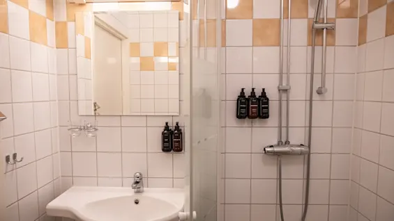 Hvitt keramisk vask med sølvkran er montert på en flislagt vegg på et bad, ledsaget av et dusjhode, glassvegg og flasker med toalettartikler. Beige fliser fremhever innredningen.
