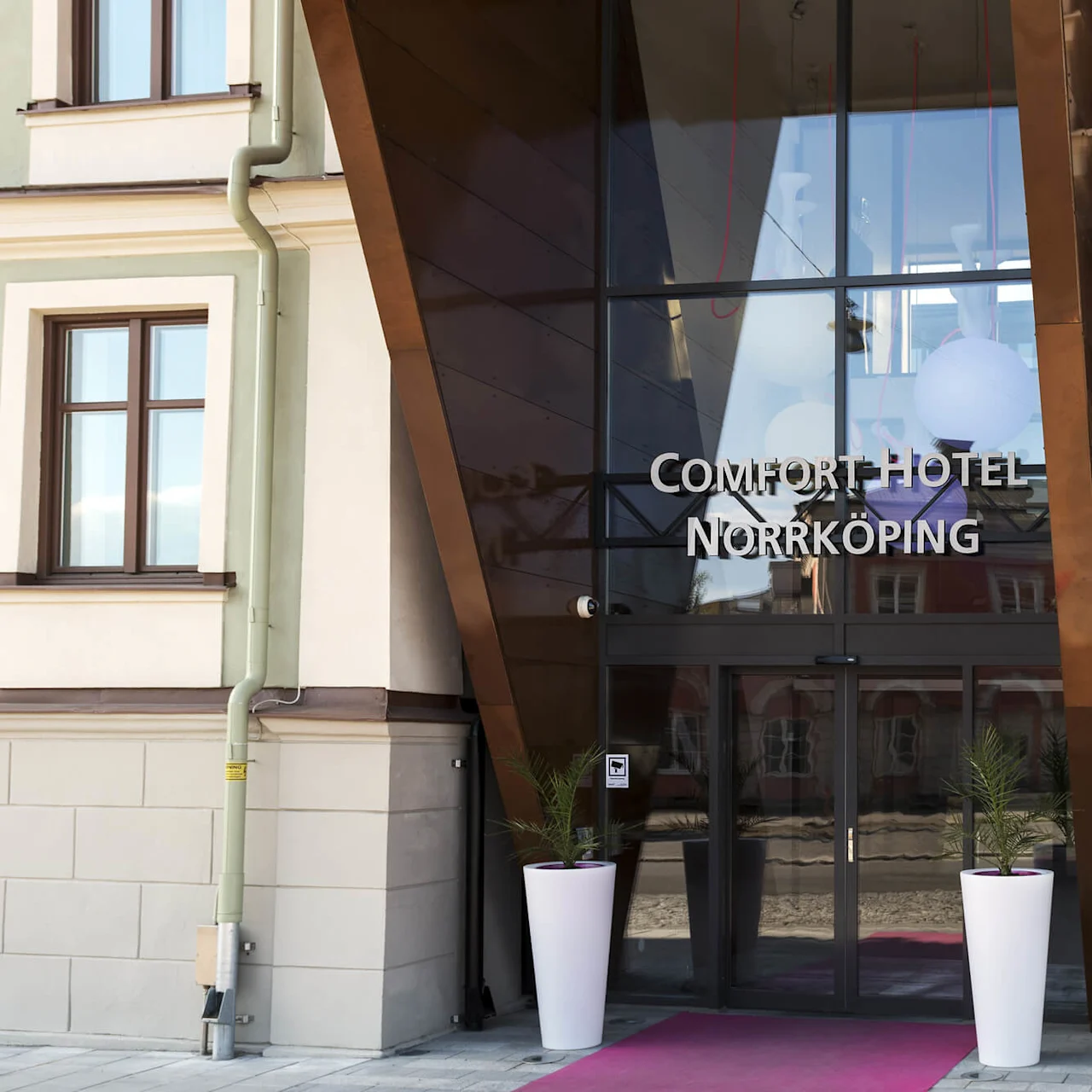 Glassdører fører til "Comfort Hotel Norrköping", flankert av planter og tradisjonell arkitektur.