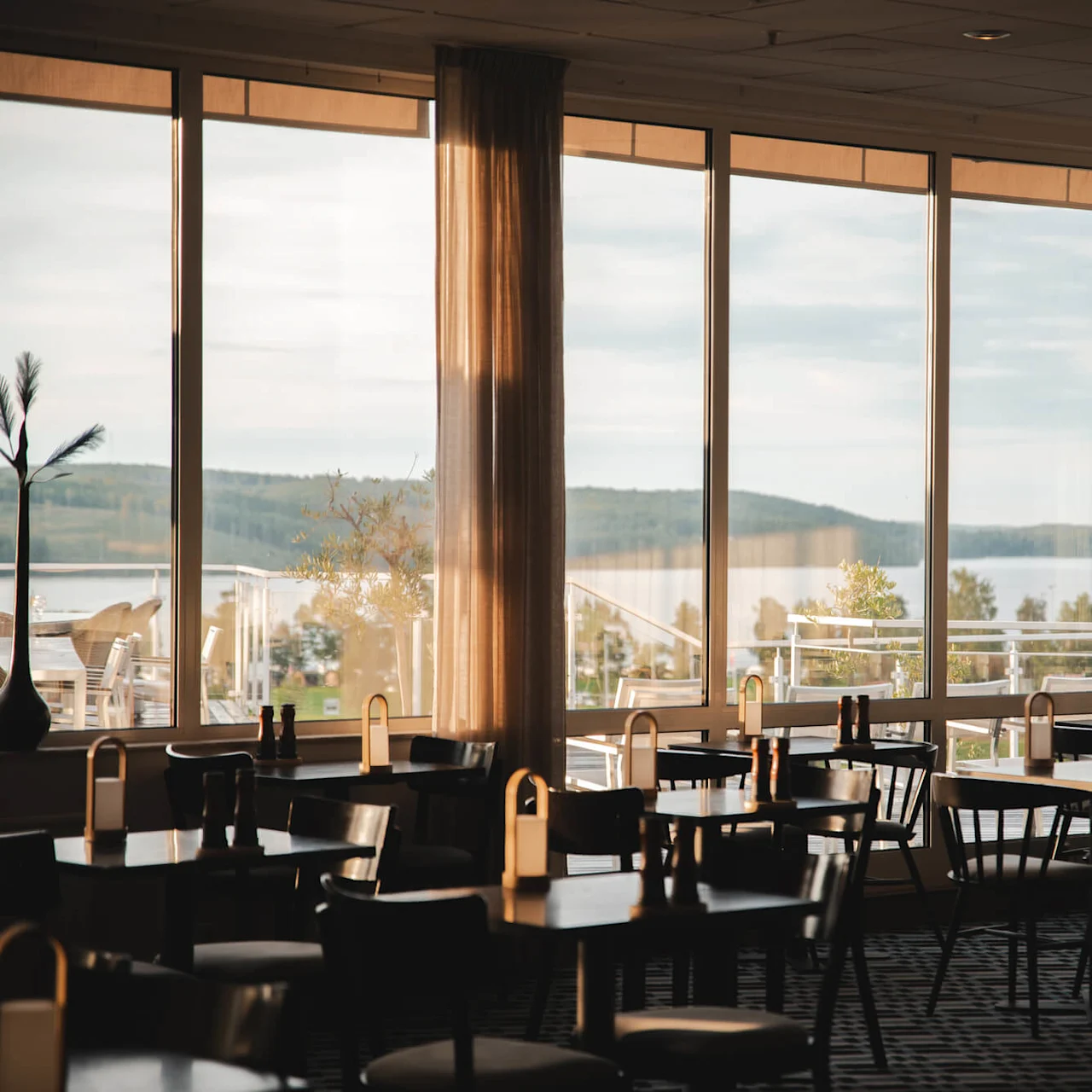 Bord og stoler er ordnet i en svakt opplyst restaurant, sollys som filtreres gjennom store vinduer med utsikt over et rolig landskap av vann og åser.