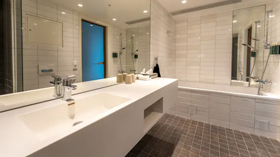 Moderne badeværelse med hvid håndvask med to haner, stort spejl, håndklæder og badekar. Flisebelagte vægge og gulve, loftlys og en trædør skaber en elegant, indbydende atmosfære.