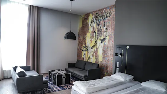 Gray couch and bed in modern room; colorful wall mural with text "The service" behind couch; large window with curtains, black lamp above table, wooden floor.
