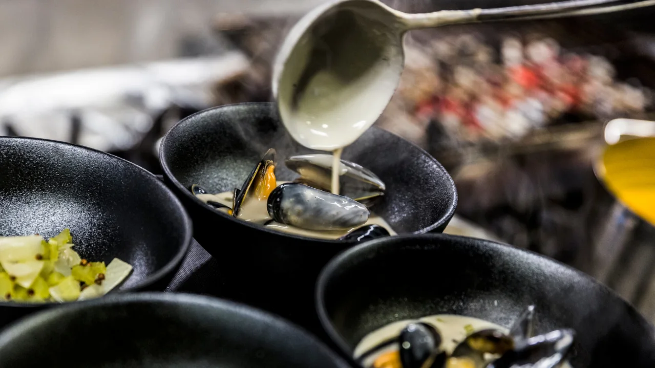 A ladle pours liquid into a pan containing mussels and vegetables in a kitchen.