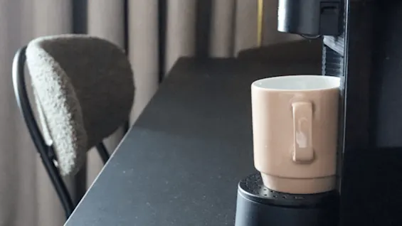 A coffee mug rests on a black coffee machine in an indoor setting, next to a chair and a yellow lamp, with grey curtains in the background.