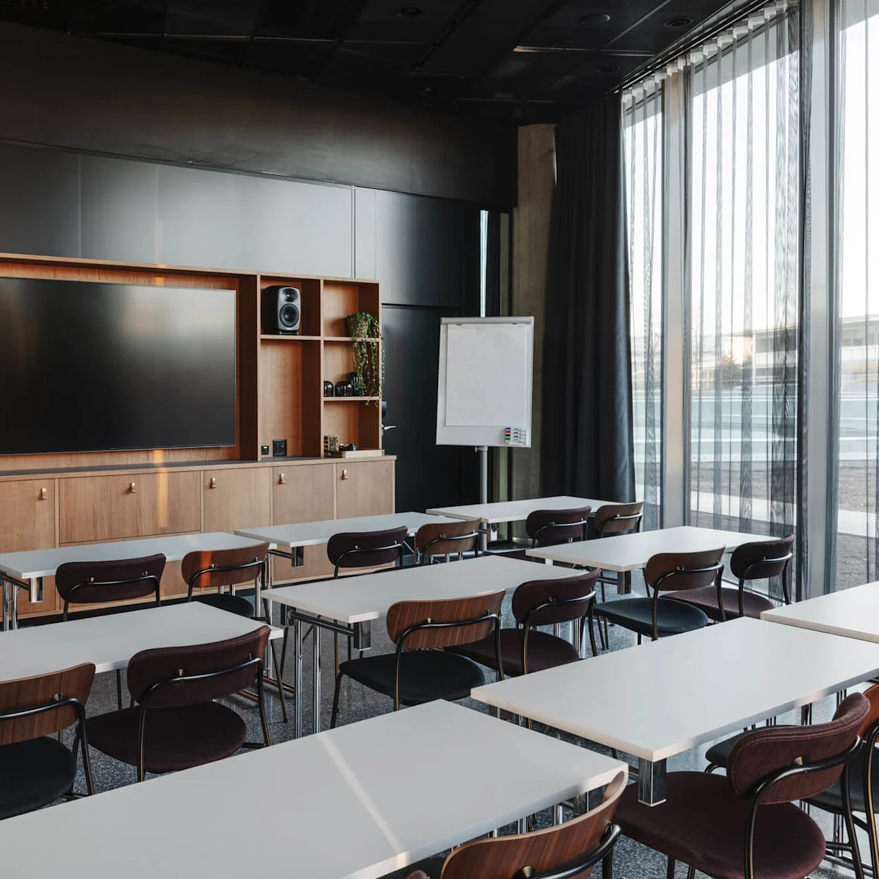 Classroom setup with rows of white desks and brown chairs facing a wooden media unit holding a large flat-screen TV. A flip chart stands by the window with sheer curtains.