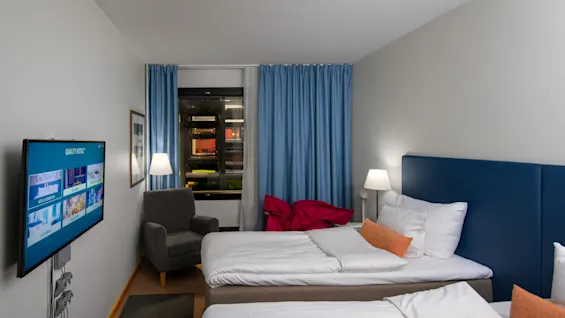 A hotel room with two neatly made beds, a wall-mounted TV displaying a welcome screen, an armchair, and a window covered by blue curtains. 
