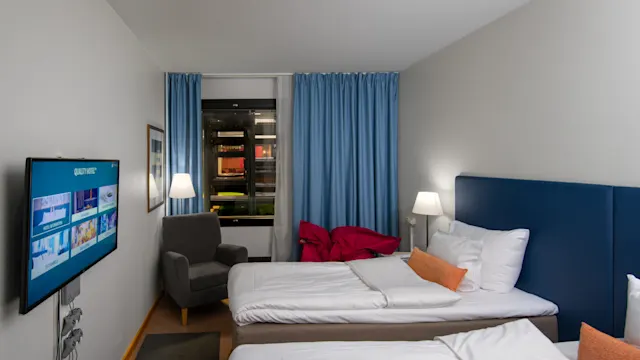 Hotellihuone, jossa on kaksi siististi pedattua sänkyä, seinälle kiinnitetty TV tervetulonäytöllä, nojatuoli ja siniset verhot ikkunassa.