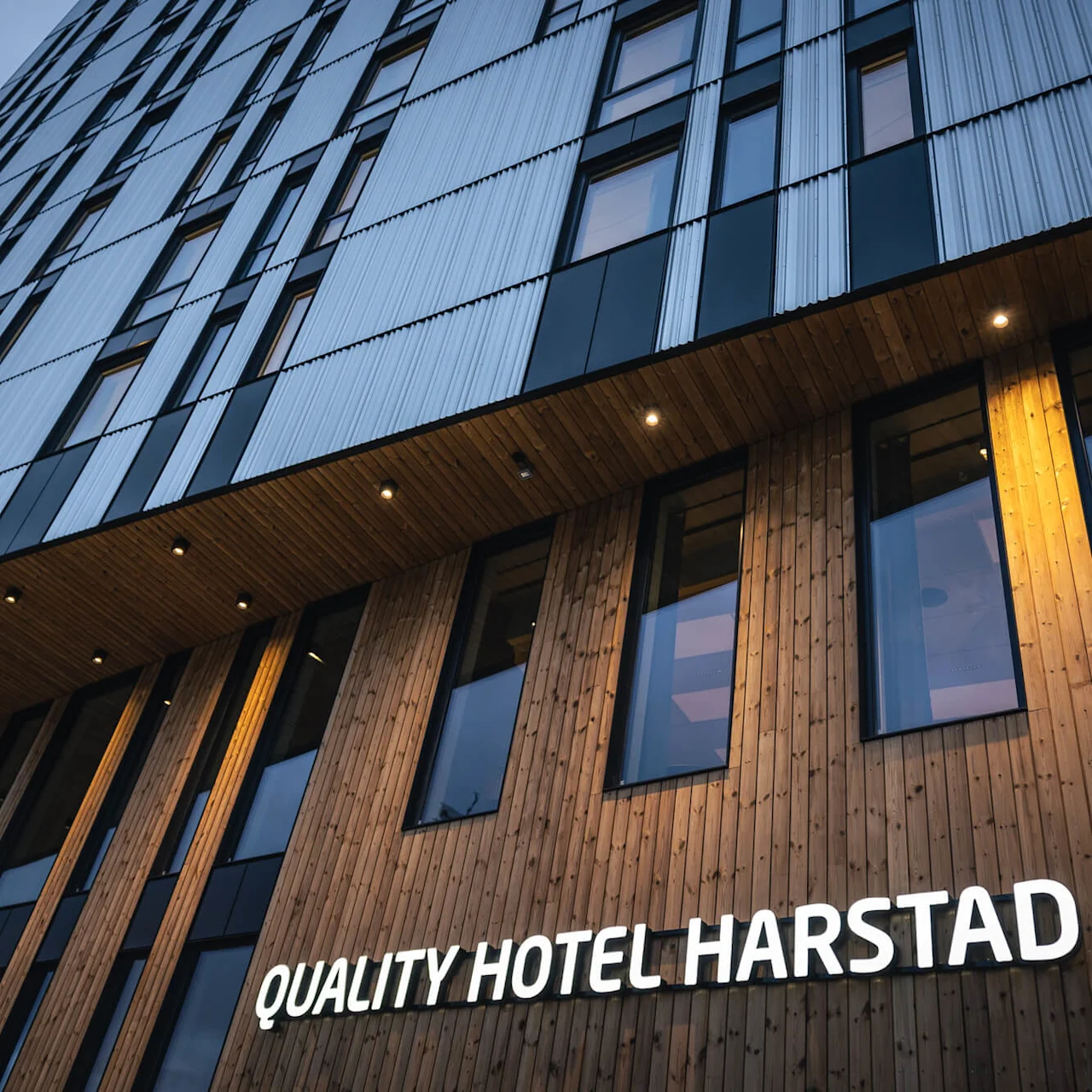 En høj bygning med træ- og metalfacade viser skiltet "QUALITY HOTEL HARSTAD" oplyst. Set nedefra om aftenen, fremhæver moderne arkitektur.