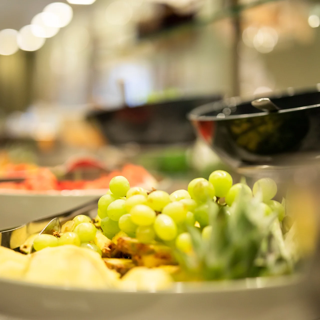 A plate of green grapes and various fruits rests on a buffet table, surrounded by other dishes, in a softly lit indoor setting.