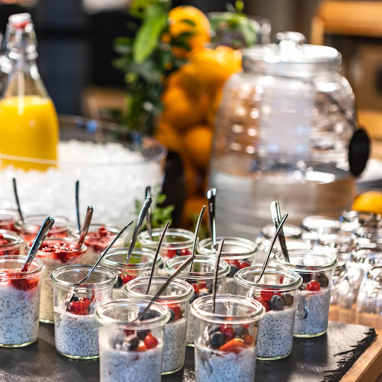 Glasses filled with chia pudding and berries are arranged on a tray, accompanied by spoons. In the background, there's a glass bottle of orange juice in ice and a container of water.