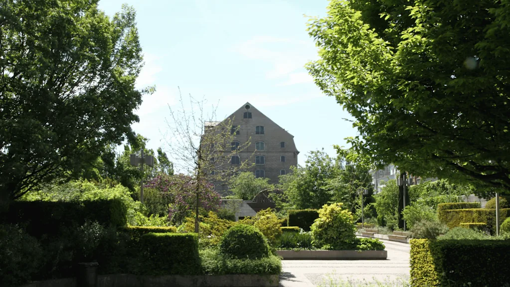 Ett stort tegelhus står bland frodig grönska, omgiven av träd, buskar och en gångväg, under en klarblå himmel.