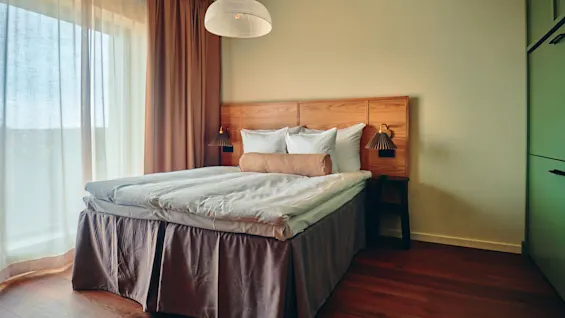 Et koselig hotellrom med en komfortabel seng, sengegavl i tre og varm belysning, badet i naturlig lys fra et stort vindu.