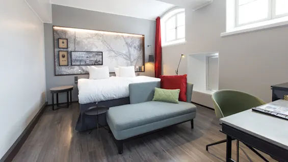 A modern bedroom with a large bed, art on the wall, a chaise lounge, and contemporary furniture in a well-lit space with large windows.
