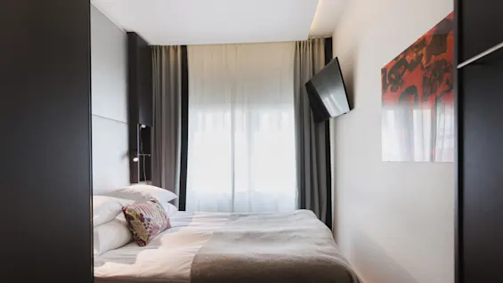 En pænt redt seng med mønstrede puder og tæppe står overfor et vægmonteret TV i et hotelværelse. Gardiner dækker vinduet, med moderne kunst på væggen.