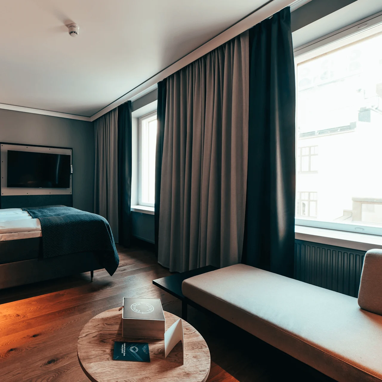 Et ryddig hotellrom med seng, bord, vegghengt TV og sofa. Naturlig lys strømmer inn gjennom to store vinduer med tykke gardiner. En brosjyre ligger på bordet.