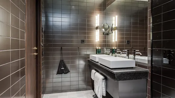 Et moderne badeværelse har to firkantede håndvaske på en sort bordplade, med oplyste spejle ovenover. Brune fliser skaber en elegant atmosfære. En pose mærket "HAIR DRYER" hænger på siden.