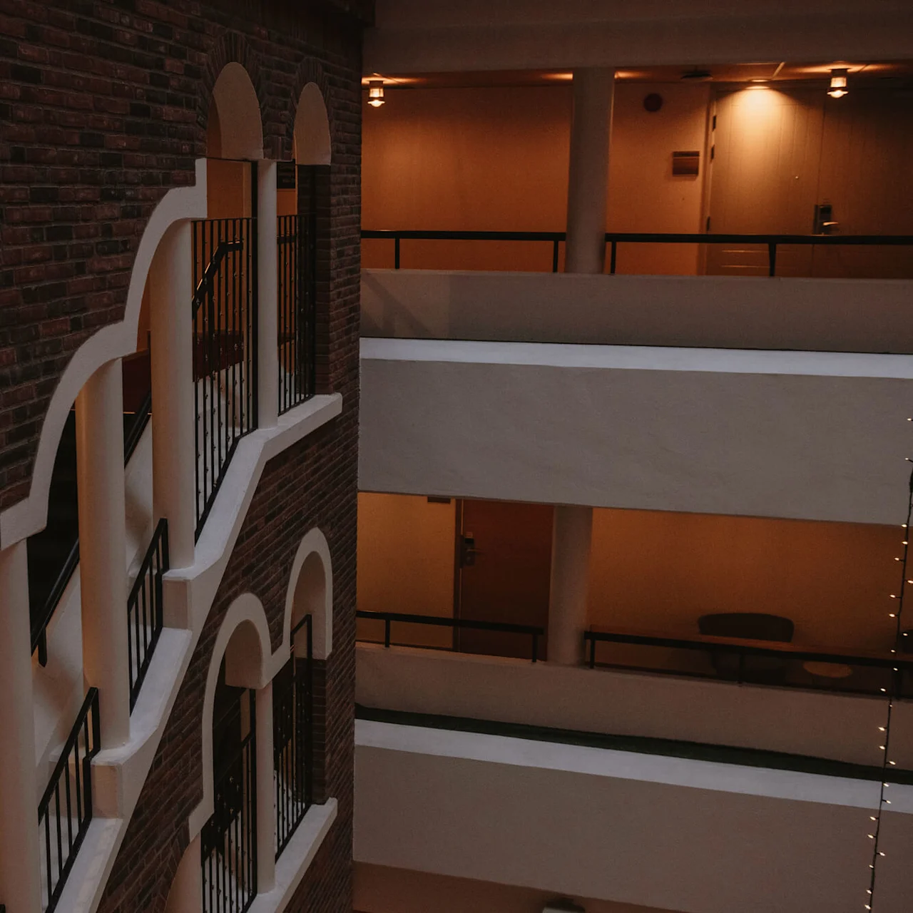 Brick wall features arched niches and balconies with black railings, facing an indoor atrium with warm lighting on multiple floors.