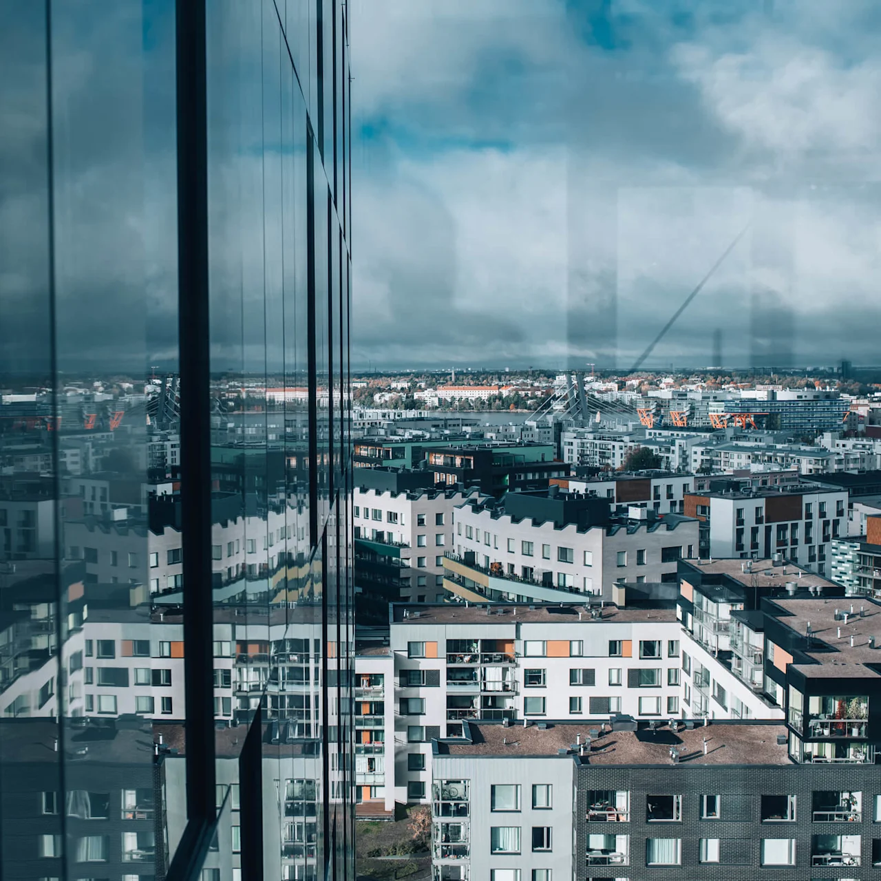 Vinduet reflekterer byens bygninger, med en overskyet himmel ovenfor. En bysilhuet med moderne lejligheder strækker sig mod horisonten under en blanding af blå himmel og mørke skyer.
