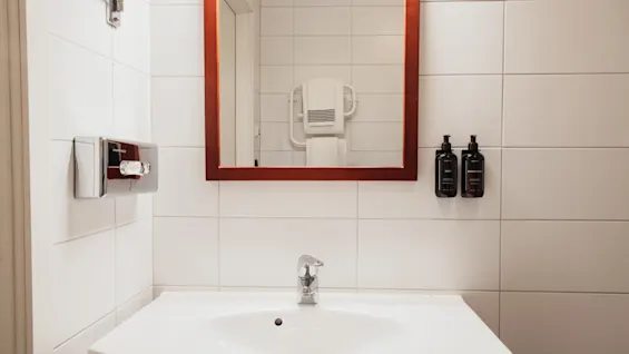 A clean and modern bathroom sink area with a mirror, soap dispenser, and toiletries, add Home Hotel Kung Oscar