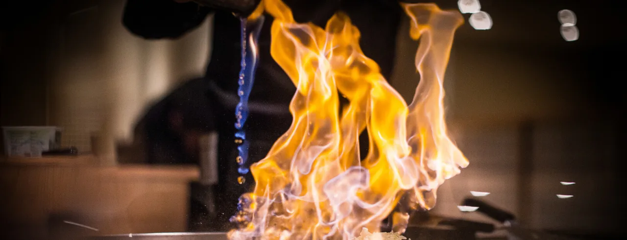 Flammor omsluter mat som tillagas på en plattgrill, kockens händer synliga, i en svagt belyst restaurangmiljö.